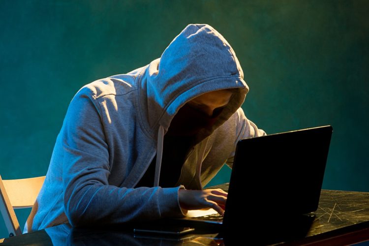 Hooded computer hacker stealing information with laptop on colored studio background